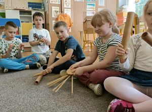 Piątka dzieci siedzi na dywanie i gra na drewnianych instrumentach.