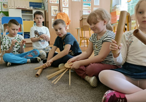 Piątka dzieci siedzi na dywanie i gra na drewnianych instrumentach.