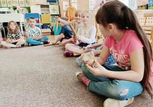 Na pierwszym planie dziewczynka w różowej koszulce gra na drewnianym instrumencie. W tle siedzą dzieci.