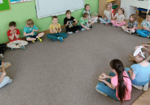 Dzieci siedzą w kole na szarym dywanie i grają na instrumentach.