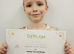 Chłopiec pozuje do zdjęcia na tle białej tablicy z dyplomem w pszczółki.