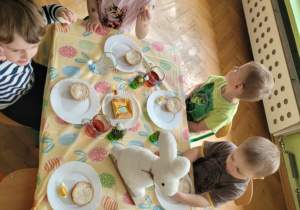 Dzieci jedzą śniadanie. Na stole stoi biały zajączek.