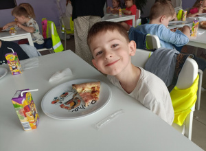 Chłopiec w białej bluzce uśmiecha się do zdjęcia. Na stole stoi talerzyk z pizzą oraz soczek w kartoniku.