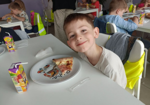 Chłopiec w białej bluzce uśmiecha się do zdjęcia. Na stole stoi talerzyk z pizzą oraz soczek w kartoniku.