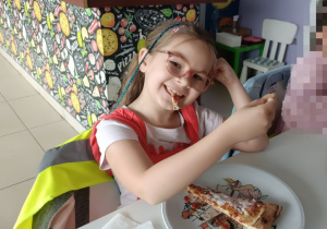 Dziewczynka w okularach zjada pizzę. Na oparciu krzesła wisi kamizelka. W tle bar w pizzerii.