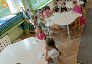 Dzieci siedzą przy czterech stolikach i jedzą babeczki oraz owoce.