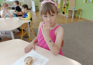 Dziewczynka w różowej bluzce i opasce siedzi przy stoliku i uśmiecha się do zdjęcia. Na stoliku leży babeczka na białej serwetce.