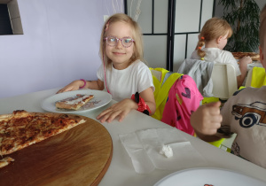 Dziewczynka w białej bluzce i okularach uśmiecha się do zdjęcia. Na stoliku stoi talerzyk z kawałkiem pizzy.