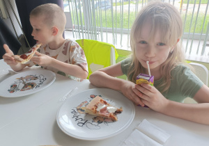 Dziewczynka pije soczek, a chłopiec siedzi obok i je pizzę.