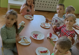 Dzieci siedzą przy stoliku i przygotowują poczęstunek. W tle stoi biały stolik z kolorowymi kubkami.
