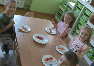 Dzieci siedzą przy stoliku i przygotowują poczęstunek. W tle widać białą szafkę z zielonymi szufladami.