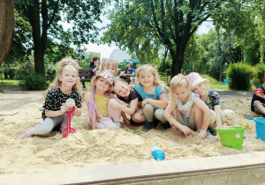 Siedem dziewczynek siedzi w piaskownicy. Dziewczynka w opasce i czarnej koszulce trzyma w ręce czerwoną łopatkę. Dwie dziewczynki mają na głowach kapelusze.
