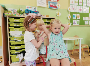 Jedna dziewczynka siedzi na krzesełku. Dziewczynka siedząca za nią przypina do włosów spinkę. Z lewej strony stoi dziewczynka w różowej opasce.