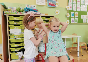Jedna dziewczynka siedzi na krzesełku. Dziewczynka siedząca za nią przypina do włosów spinkę. Z lewej strony stoi dziewczynka w różowej opasce.