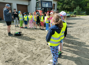 Dzieci stoją w kamizelkach odblaskowych w półkolu. Pani prezentuje przedmioty.