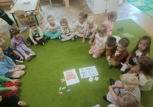 Dzieci siedzą w kole na zielonym dywanie. W środku leżą karty demonstracyjne.