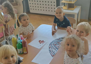 Piątka dzieci siedzi przy stoliku i maluje kropkową pracę farbami. Na stoliku stoi pięć butelek farb.