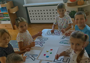 Szóstka dzieci maluje farbami napis Dzień Kropki. Wszyscy siedzą przy stoliku.