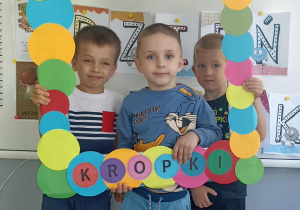 Trzech chłopców stoi na tle białej tablicy z wykonaną pracą plastyczną.
