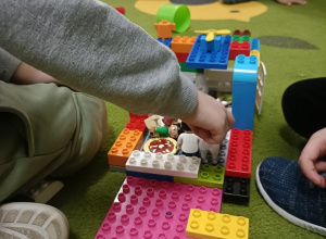 Na zielonym dywanie stoi budowla z klocków lego DUPLO. Chłopiec w szarej bluzce łączy ze sobą elementy.
