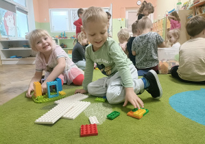 chłopiec w zielonej i dziewczynka w różowej bluzce siedzą na zielonym dywanie i bluzce buduje budowlę z klocków