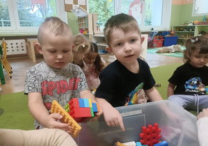 Chłopiec w szarej koszulce wyjmuje klocki z pudełka, obok siedzi chłopiec w czarnej koszulce