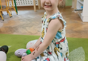 Dziewczynka w jasnej sukience siedzi na dywanie, uśmiecha się do zdjęcia, a w ręce trzyma zwierzątko.