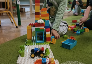 Na zielonym dywanie stoi budowla z klocków lego DUPLO. Chłopiec w zielonej bluzce łączy ze sobą elementy.