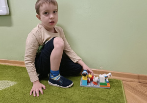 Chłopiec siedzi na tle ściany. Na dywanie stoi budowla z klocków LEGO DUPLO.