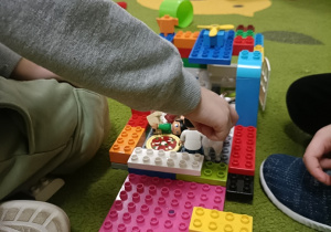 Na zielonym dywanie stoi budowla z klocków lego DUPLO. Chłopiec w szarej bluzce łączy ze sobą elementy.