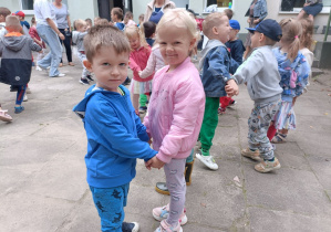 Chłopiec i dziewczynka trzymają się za ręce. Dziewczynka ma na sobie różową kurtkę a chłopiec niebieską bluzę. W tle widać grupę dzieci.