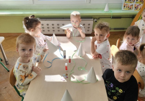 Piątka dzieci siedzi przy stoliku na którym stoją białe czapki, kleje i papierowe kropki.