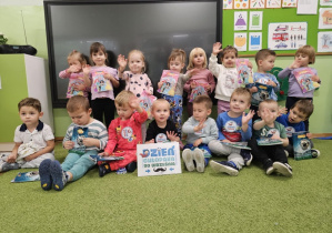 Dzieci siedzą na zielonym dywanie i machają do zdjęcia. Chłopiec w czarnej bluzce trzyma kartkę z napisem "Dzień Chłopaka"