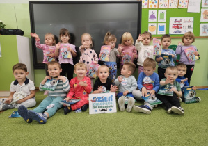 Dzieci siedzą na zielonym dywanie i machają do zdjęcia. Chłopiec w czarnej bluzce trzyma kartkę z napisem "Dzień Chłopaka"