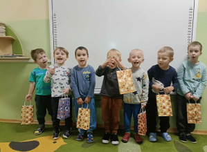 Chłopcy stoją na zielonym dywanie. W rękach trzymają torby z prezentami. W tle biała tablica.