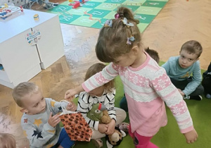 Dziewczynka w różowym sweterku wręcza prezent koledze, który siedzi na dywanie.