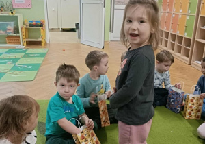 Dziewczynka w ciemnej bluzce wręcza prezent koledze, który siedzi na dywanie.