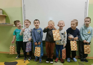 Chłopcy stoją na zielonym dywanie. W rękach trzymają torby z prezentami. W tle biała tablica.