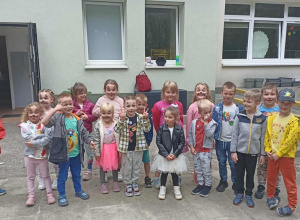 Grupa dzieci stoi na chodniku. W tle widać budynek przedszkola oraz otwarte drzwi.