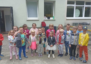 Grupa dzieci stoi na chodniku. W tle widać budynek przedszkola oraz otwarte drzwi.