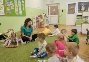Grupa dzieci siedzi z panią prowadzącą na dywanie i wykonuje ćwiczenia.
