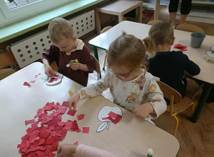 Chłopiec i dziewczynka w okularach siedzą przy stoliku i przyklejają kartki papieru na szablon jabłka.