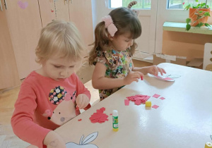 Dwie dziewczynki siedzą przy stoliku i przyklejają kartki papieru na szablon jabłka.