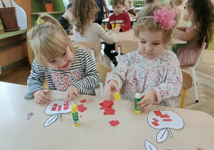 Dwie dziewczynki siedzą przy stoliku i przyklejają kartki papieru na szablon jabłka.