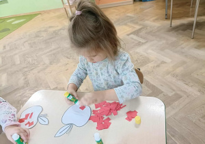 Dziewczynka w kitce siedzi przy stoliku i przykleja kartki papieru na szablon jabłka.
