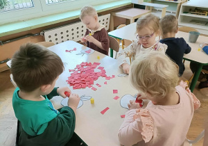 Czwórka dzieci siedzi przy stoliku i przykleja kartki papieru na szablon jabłka.