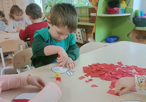 Chłopiec w zielonej bluzie siedzi przy stoliku i przykleja kartki papieru na szablon jabłka.