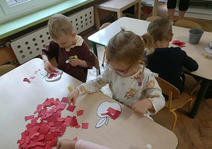 Chłopiec i dziewczynka w okularach siedzą przy stoliku i przyklejają kartki papieru na szablon jabłka.