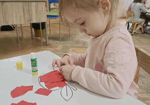 Dziewczynka w różowej bluzce siedzi przy stoliku i przykleja kartki papieru na szablon jabłka.
