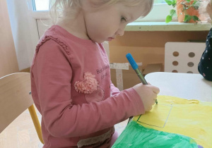 Dziewczynka w różowej bluzce siedzi na krzesełku, w dłoni trzyma niebieski mazak i rysuje po śladzie.